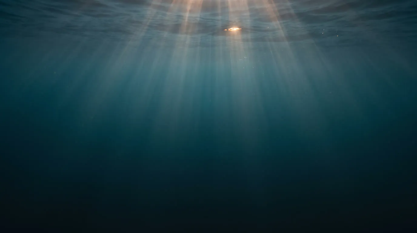 Lichtstrahlen fallen von der Oberfläche schräg in dunkles, tiefblaues Wasser, oben ein kleiner warmer Lichtpunkt.