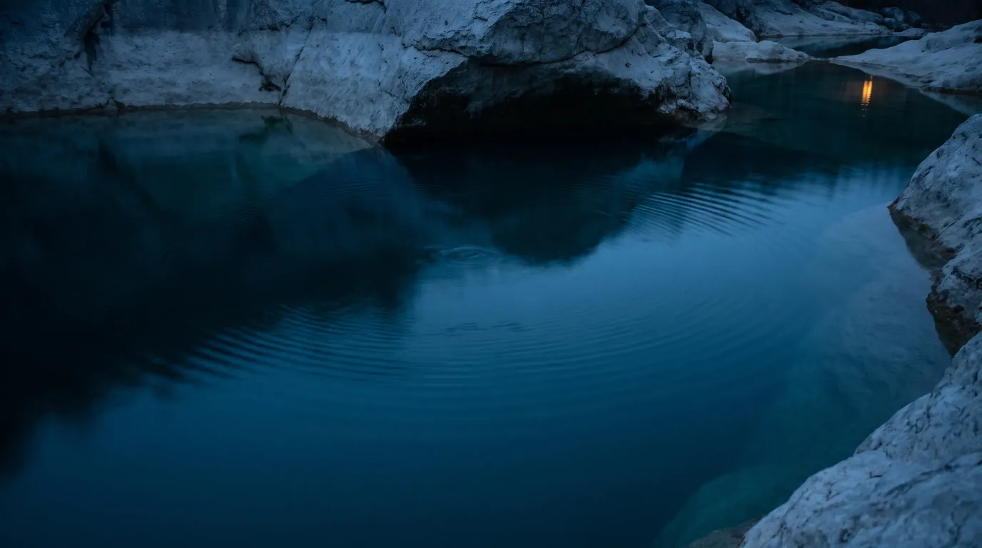 Eine stille Quelle in einer dunklen Felsmulde, das Wasser tiefblau, am Rand ein schmaler warmer Lichtschein.